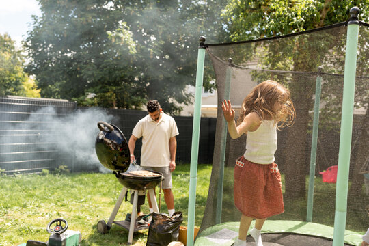 Dad is cooking BBQ, while his daughter is jumping on a trampoline