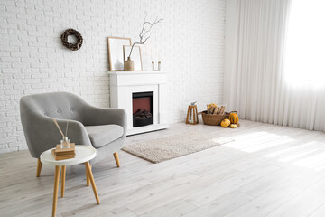 Naklejka premium Interior of stylish room with modern fireplace, armchair and autumn decorations near white brick wall