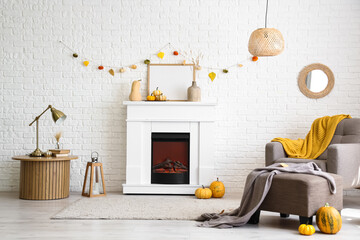 Interior of stylish room with modern fireplace, armchair, table and autumn decorations near white brick wall