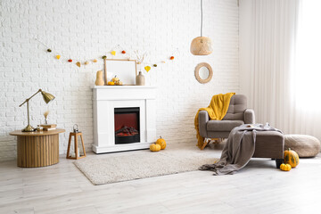 Interior of stylish room with modern fireplace, armchair, table and autumn decorations near white brick wall