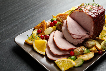 Tasty baked ham served with vegetables and oranges on black table