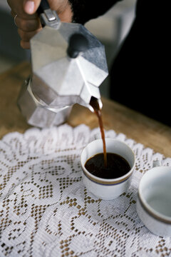 Hand pouring coffee into cup from moka pot on lace mat.