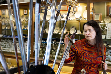 Positive woman traveler viewing with interest at various handmade forged swords in gift shop of...