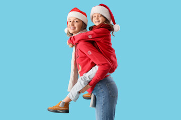 Fototapeta premium Beautiful young woman and her cute little daughter in Santa hats on blue background. Christmas celebration