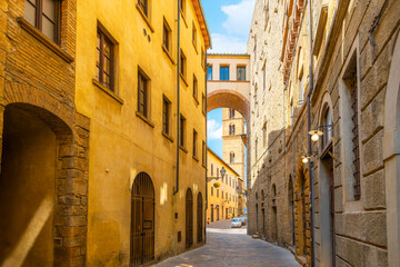 A picturesque narrow alley towards the Volterra Duomo Cathedral bell tower of Torre Campanaria, and the Buonparenti House Tower in the medieval old town of the Tuscan hill town of Volterra, Italy.