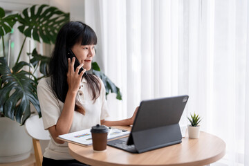 Asian businesswoman talking on phone and using tablet while working in modern office