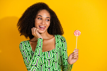 Charming young woman with curly hair wearing a green blouse holding a lollipop against a vibrant yellow background.