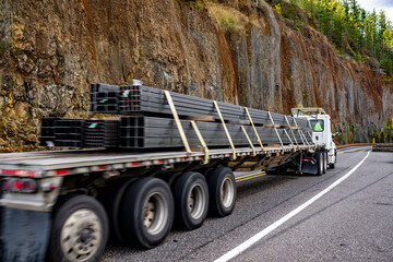 Day cab white big rig semi truck transporting heavy fastened metal cargo on flat bed semi trailer running on narrow mountain road with rock wall on the side