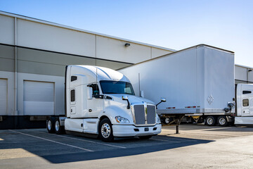 Powerful white big rig semi truck tractor standing side by side with loading cargo dry van semi...