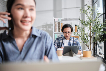 Asian Japanese male startup worker concentrates on working with digital tablet at desk near daylight window in casual small business office, online freelance jobs, and female colleague foreground.