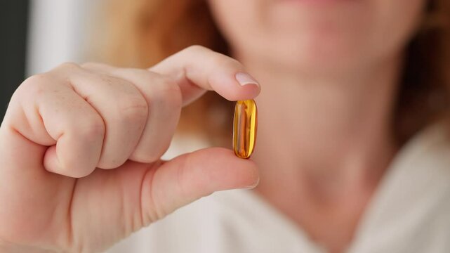 Close-up woman&rsquo;s hand holding two fingers orange capsule with Omega D3 in camera and puts it near her mouth white background smily. Health, complex vitamins, self-care people. Taking medication, pills
