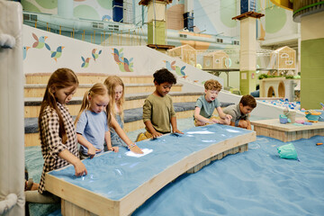Friendly little boys and girls creating drawings on long table with blue sand while spending time...