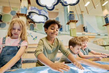 Happy little boy making pile of blue sand on tabletop between other kids drawing pictures on spacious playground