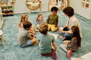 Group of cute intercultural kids sitting in circle on the floor and looking at young female teacher...