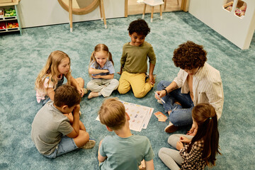 Young woman with paper cards in hand talking to little girl while sitting on the floor among intercultural kids at lesson