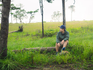 Young man in green jacket and hat resting on log amidst forest and green grass during sunset at Phu Soi Dao, Thailand.