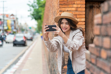 Asian Tourist Woman In Hat and Jacket Using Phone Beside A Brick Wall in Chiang Mai, Thailand