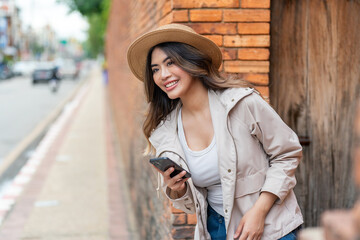 Asian Tourist Woman In Hat and Jacket Using Phone Beside A Brick Wall in Chiang Mai, Thailand