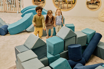 Happy little boy and two pretty girls moving towards heap of big soft toy cubes before building...