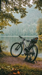 Fototapeta premium A mountain bike rests by a serene lake, surrounded by lush greenery and misty mountains.