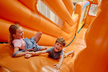 Cute little girl holding her friend by hand while both lying on top of steep orange leather slide and trying not to roll down