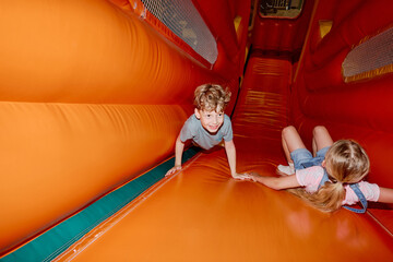 Cute little girl lying on steep soft orange leather slide and holding boy by hand while her friend...