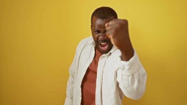 Angry african american man wearing shirt standing with fist raised, fury and frustration on isolated yellow background, mad rage expressed aggressively
