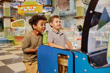Obraz premium Two cute excited intercultural boys looking at screen of slot machine and gambling while having fun at leisure center