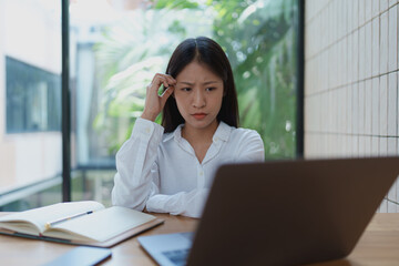 Young woman in thoughtful concentration while working on laptop at a bright indoor workspace surrounded by greenery, contemplating her next steps in a project