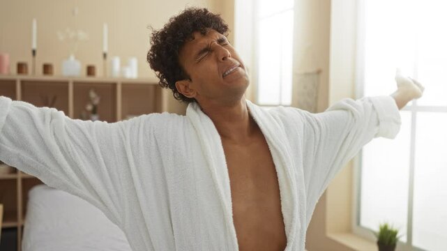 Young man yawning and stretching in a spa room with a cozy interior, wearing a white robe and expressing morning tiredness and relaxation.