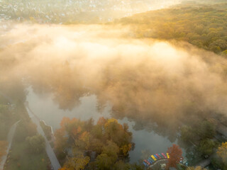 A misty autumn lake with vibrant orange, red, and green foliage surrounds the calm water. The fog adds a serene and mysterious atmosphere to the scene, with trees reflected on the still lake surface