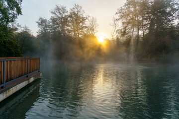 A misty autumn lake with vibrant orange, red, and green foliage surrounds the calm water. The fog adds a serene and mysterious atmosphere to the scene, with trees reflected on the still lake surface