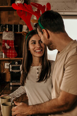 Loving couple for Christmas at home. They are standing in the kitchen, using phone, and drinking tea or coffee.