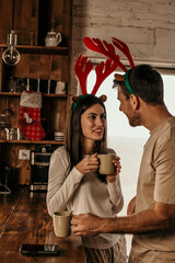 Festive couple for Christmas at home. They are standing in front of the kitchen window and drinking tea or coffee.