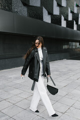 Stylish woman wearing a black leather jacket and white pants walks down a sidewalk