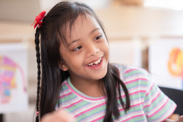 Asian joyful young girl with Down syndrome smiles warmly while engaged in a creative activity. Her cheerful expression highlights the happiness and warmth of the moment