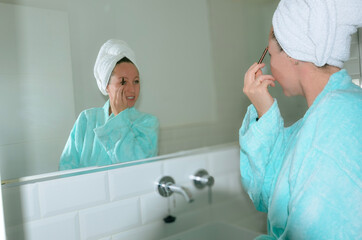 Obraz premium Woman applying makeup in bathroom mirror wearing bathrobe and towel turban