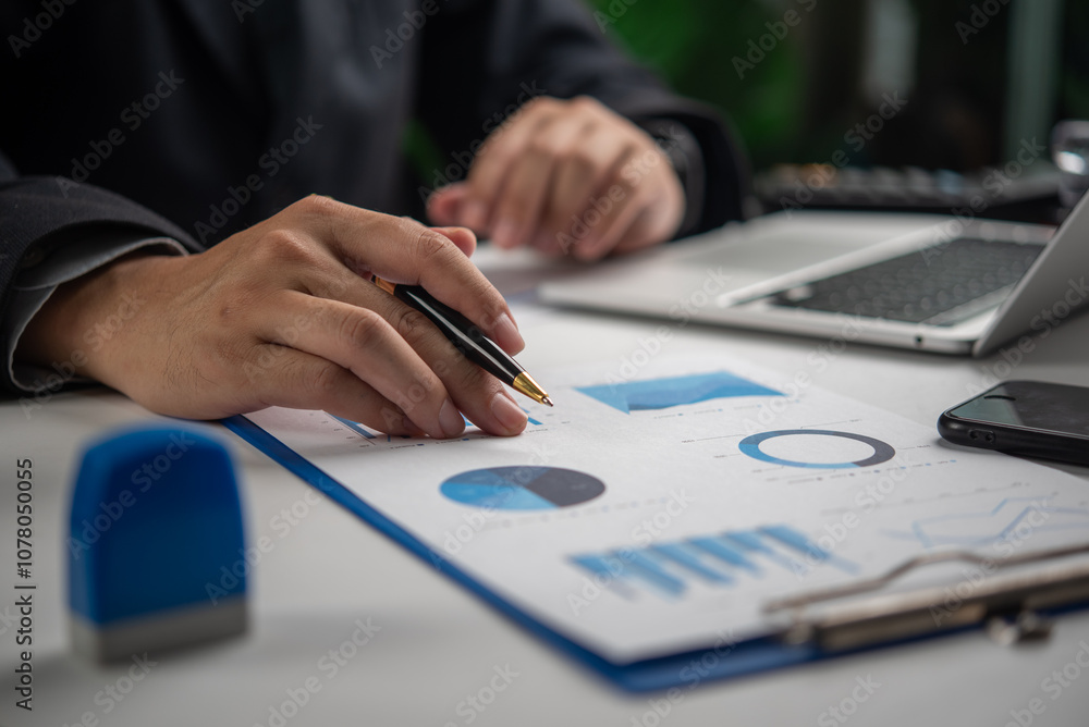 Poster a person reviews business data charts on a paper document with a pen in hand.