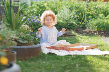 Little curly pretty girl sitting on lawn and eating delivered pizza from box in summer shiny...