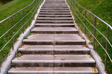 Fototapeta premium Staircase with stone steps and steel railings. Hill with green grass. Staircase in the park covered with autumn leaves.