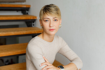 Portrait of  millennial woman with short blonde hair, in front of a white backdrop, wearing a beige...