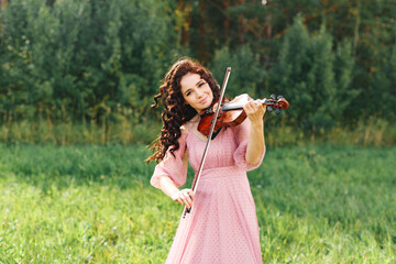 Beautiful girl with curly black hair in pink dress plays the violin in field at summer forest,