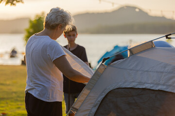 Grandparent and Grandson Setting Up a Tent by the Lake at Sunset, Capturing Family Bonding, Outdoor Adventure, and Generational Connection Amidst the Beauty of Nature in a Tranquil Setting