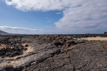 White sand beach with solidified volcanic lava flows, black rocks. Mountains in the background and sky with big white clouds. Caletón Blanco, Lanzarote, Canary Islands, Spain