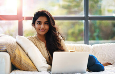 Shot of pretty young woman working with her laptop while sitting on couch at home.