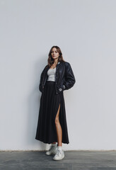 Young woman in casual chic outfit wearing black maxi skirt with slit, bomber jacket, white tank top and sneakers posing against gray studio background