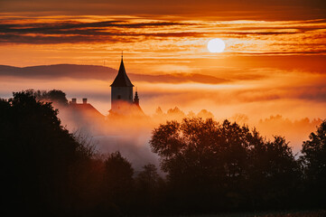 Obraz premium Silhouette of a Christian Church in a Beautiful Autumn Morning During Sunrise, Rural Landscape with Orange Sky and Soft Morning Mist Over the Village