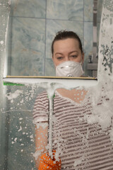 A woman cleans a shower area using a squeegee, wearing a protective mask. Effective cleaning methods.