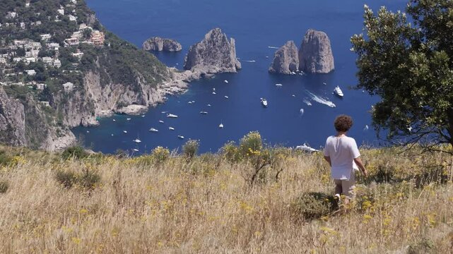 Revealing drone shot of an Italian man walking toward a tree, showing Capri&rsquo;s Faraglioni rocks and blue ocean
