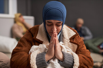 Front view of adult Middle Eastern woman praying with eyes closed in refugee shelter